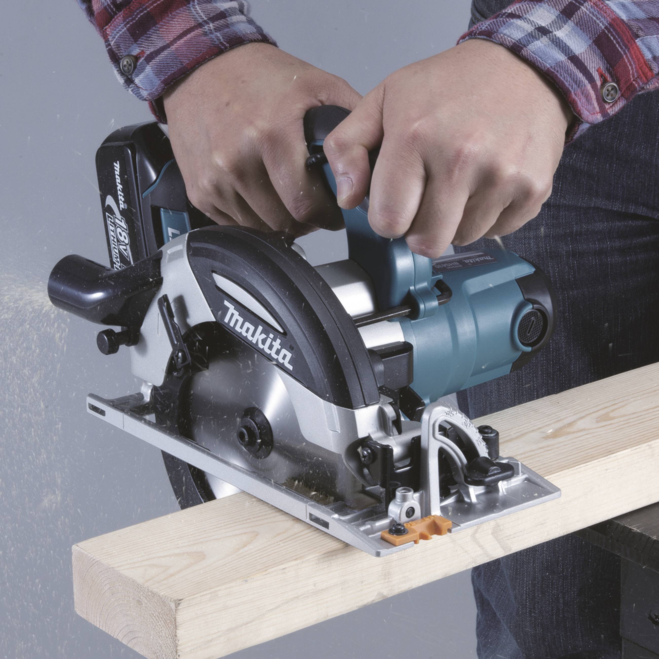 Eine Person schneidet mit einer elektrischen Handkreissäge durch ein Holzbrett. Die Säge erzeugt Späne und Staub.