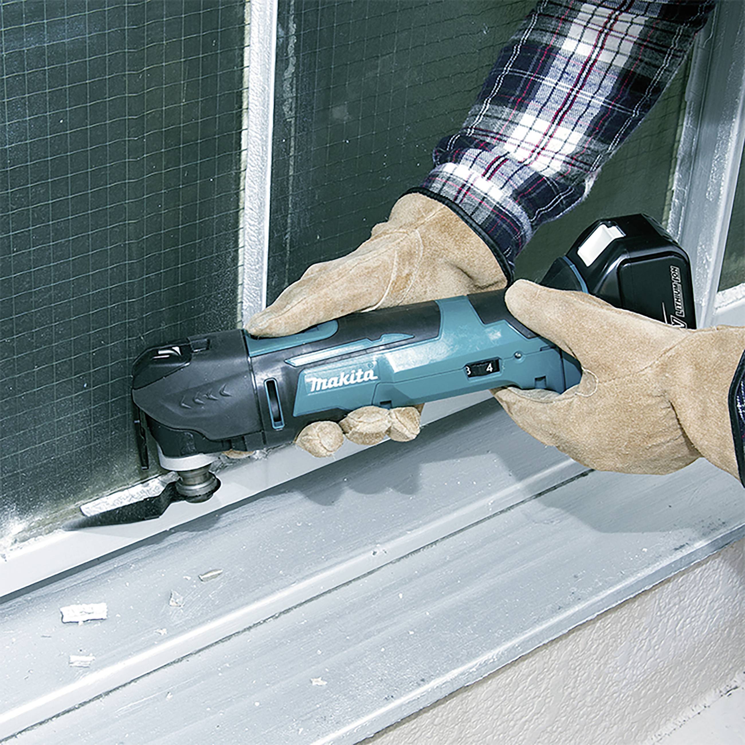 Eine Person arbeitet mit einem blauen Mehrzweckwerkzeug an einem Fensterrahmen. Sie trägt Handschuhe und ein kariertes Hemd.