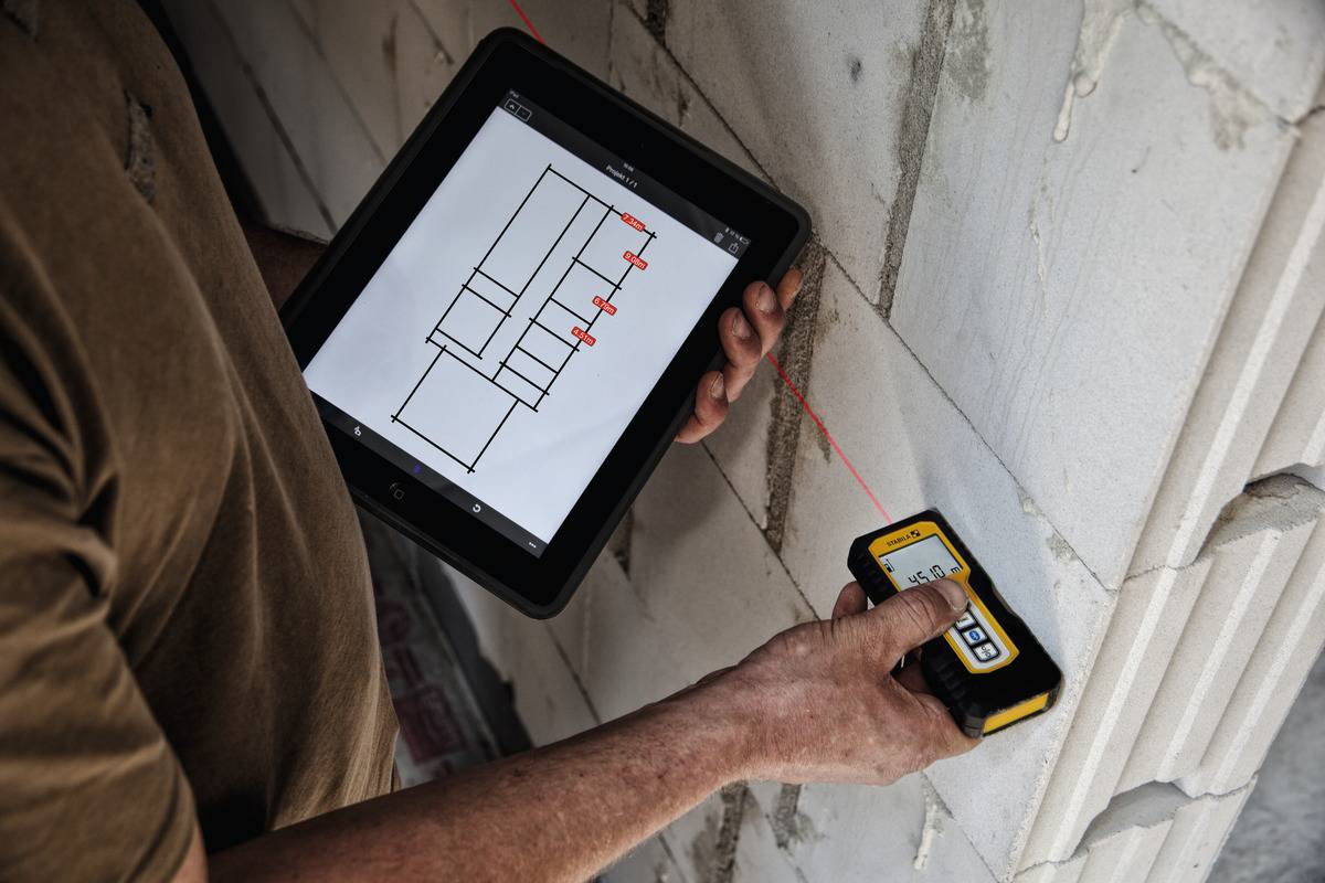 Person hält ein Tablet mit Bauplänen und misst mit einem Laserentfernungsmesser die Wand. Hauptfokus auf Präzisionsmessung.