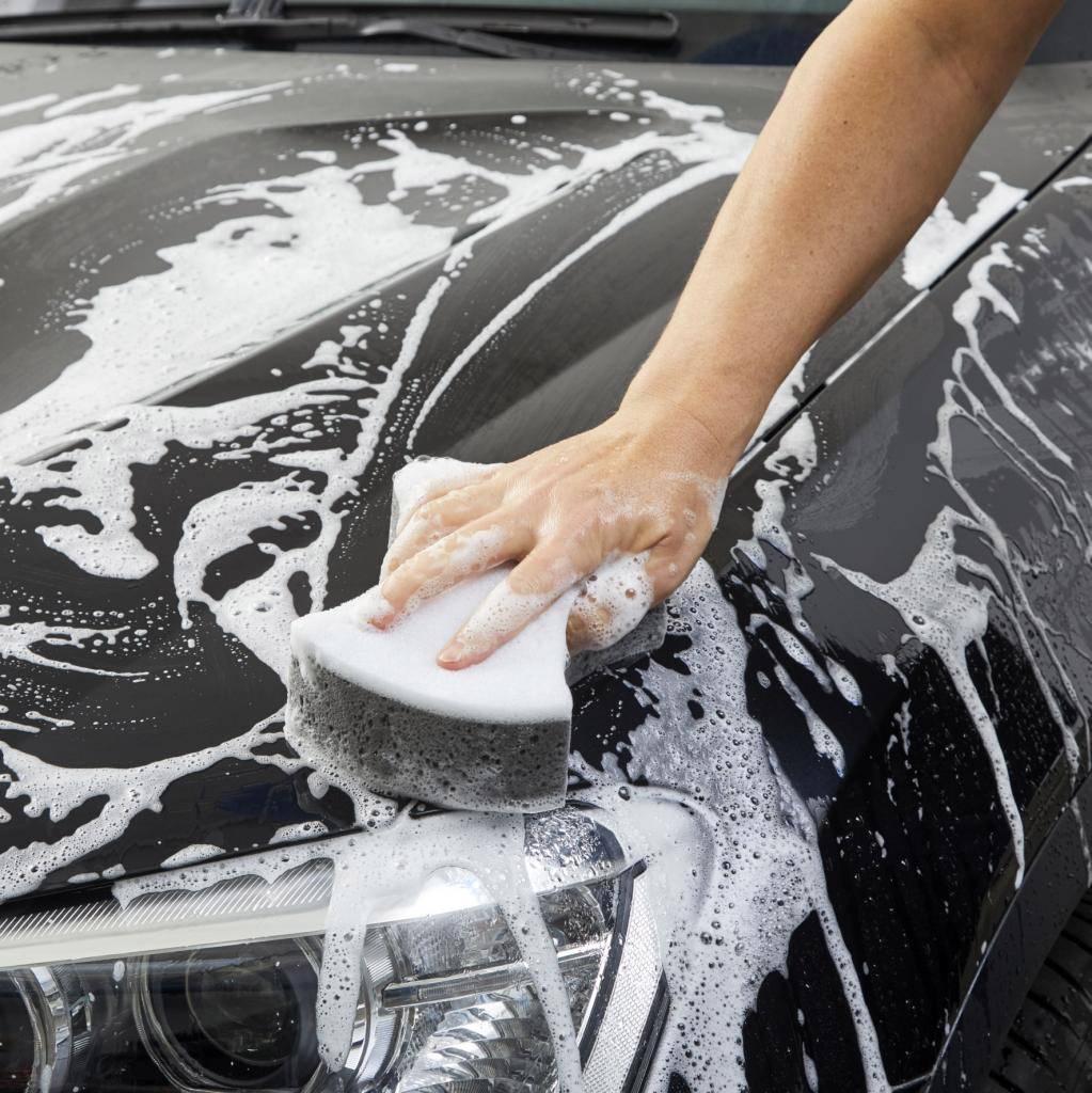 Eine Hand wäscht mit einem Schwamm ein Auto, das mit Schaum bedeckt ist. Der Fokus liegt auf der Reinigung der Motorhaube und Scheinwerfer.