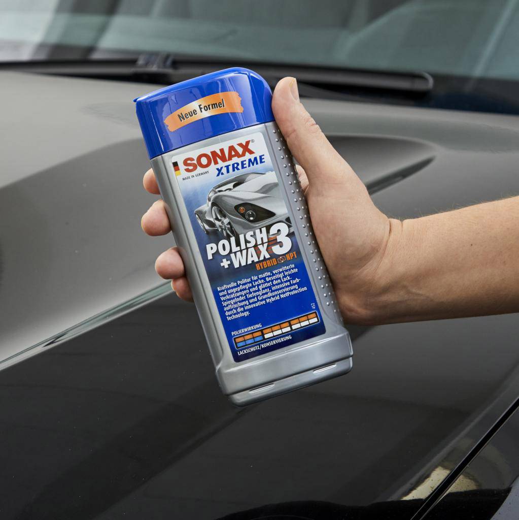 Eine Hand hält eine Flasche 'Sonax Xtreme Polish + Wax 3', ein Autopolitur- und Wachsprodukt. Der Hintergrund zeigt ein dunkles Auto.