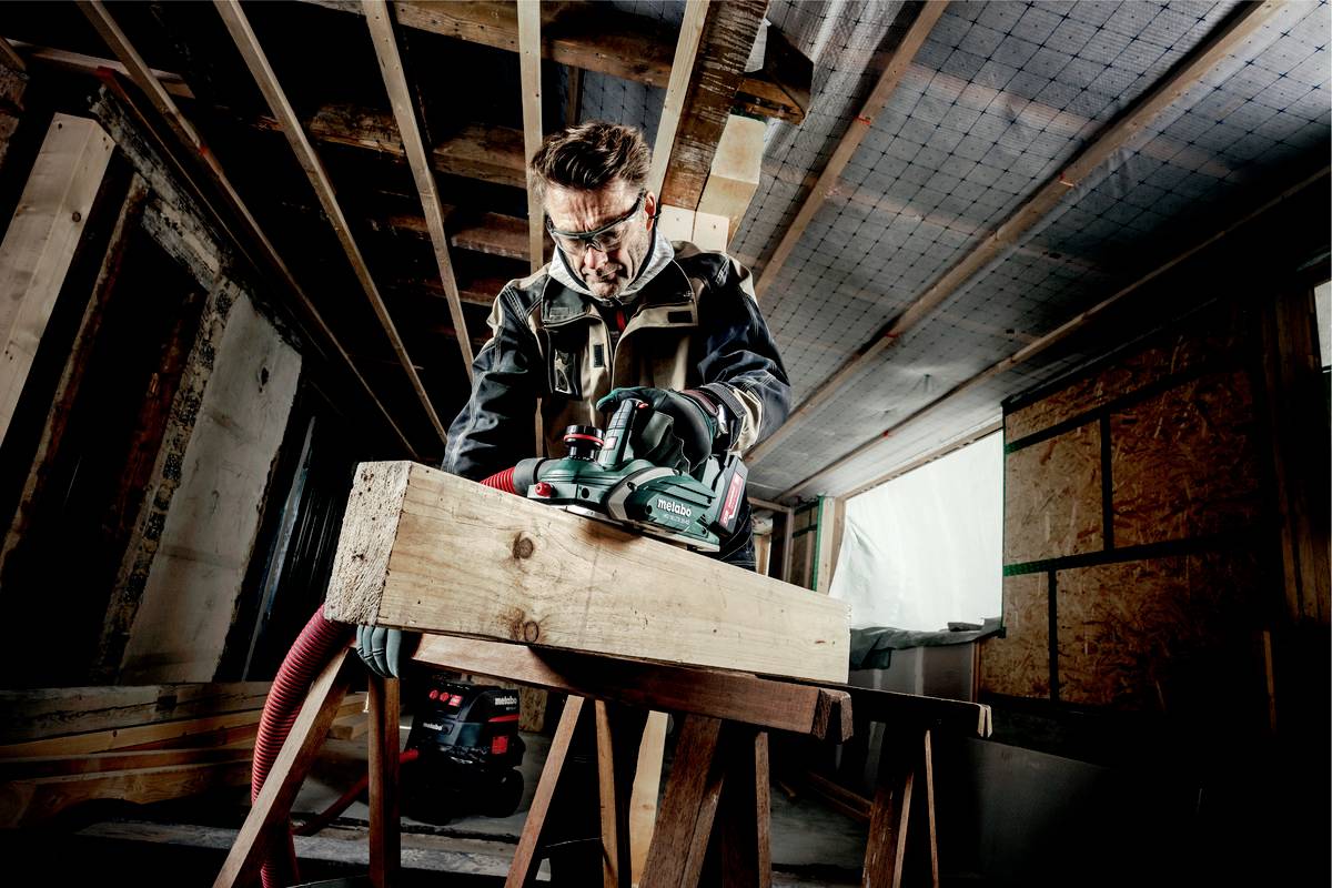 Ein Handwerker mit Schutzbrille arbeitet mit einer Elektrosäge an einem Holzstück in einem halbfertigen Raum.