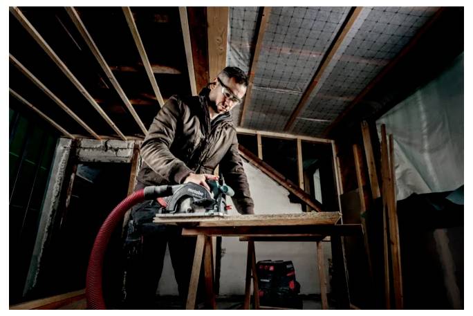 Eine Person mit Schutzbrille bedient eine Kreissäge auf einem Holzbrett in einem teilweise konstruierten Raum mit freiliegenden Balken und sichtbaren Werkzeugen.