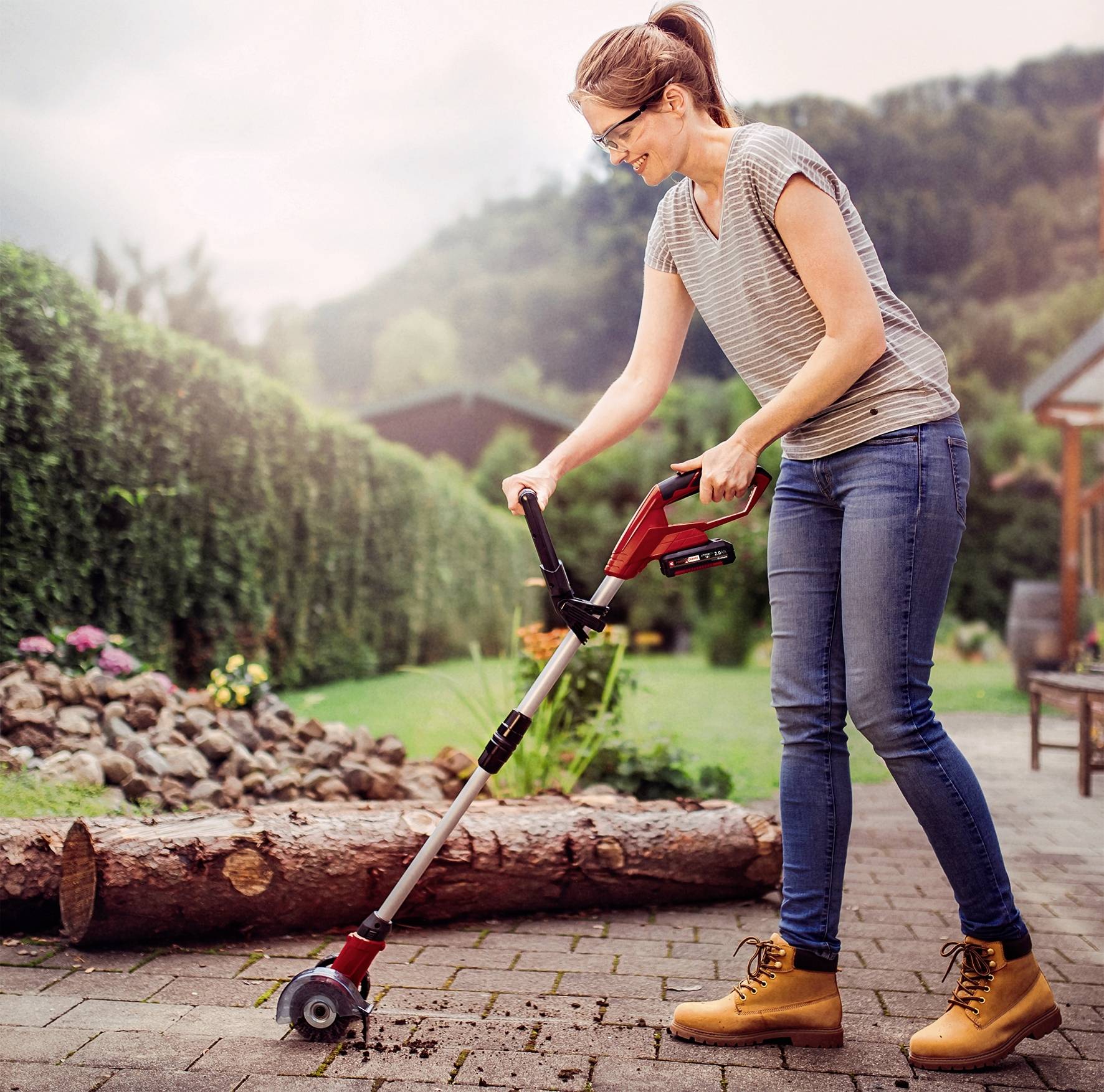 Eine Frau nutzt ein Gartenwerkzeug mit Rädern, um den Weg zwischen Beeten zu säubern, umgeben von Bäumen und einer Wiese.
