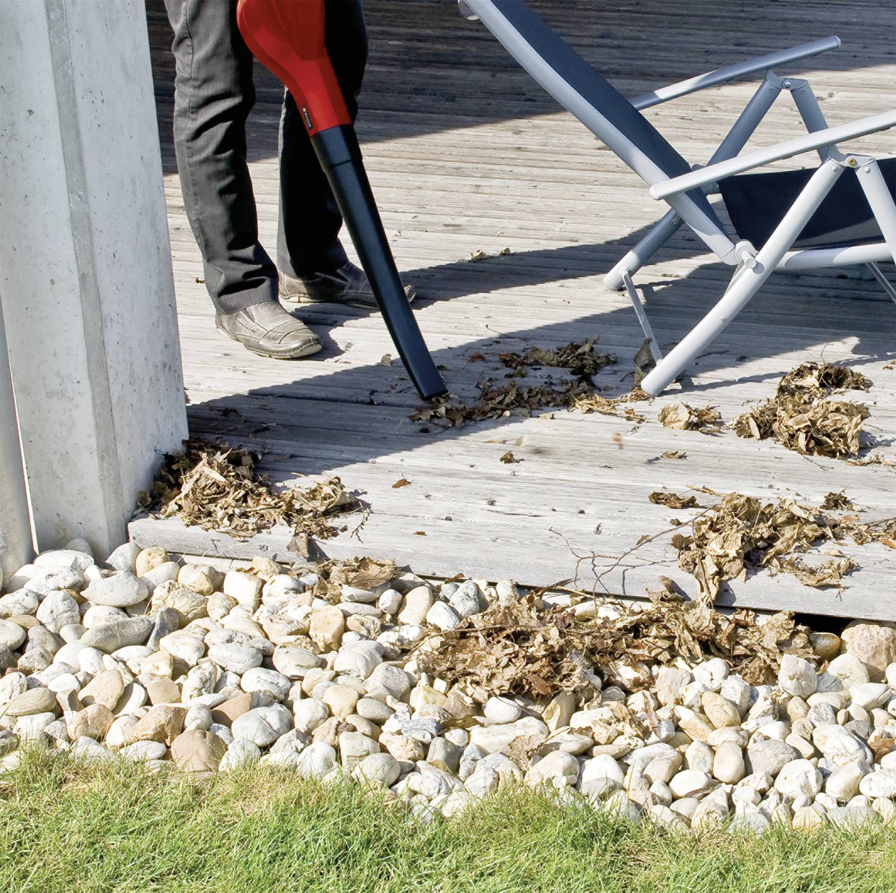 Eine Person verwendet einen Laubbläser, um Laub von einer Holzterrasse zu entfernen. Im Vordergrund sind Steine und ein zusammengeklappter Stuhl zu sehen.