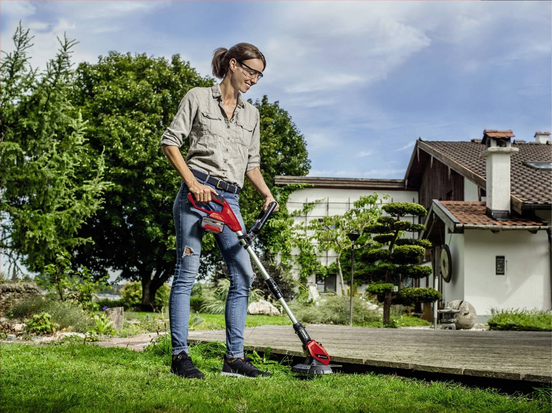 Eine Frau mäht den Rasen mit einem elektrischen Rasentrimmer in einem gepflegten Garten vor einem Haus bei sonnigem Wetter.