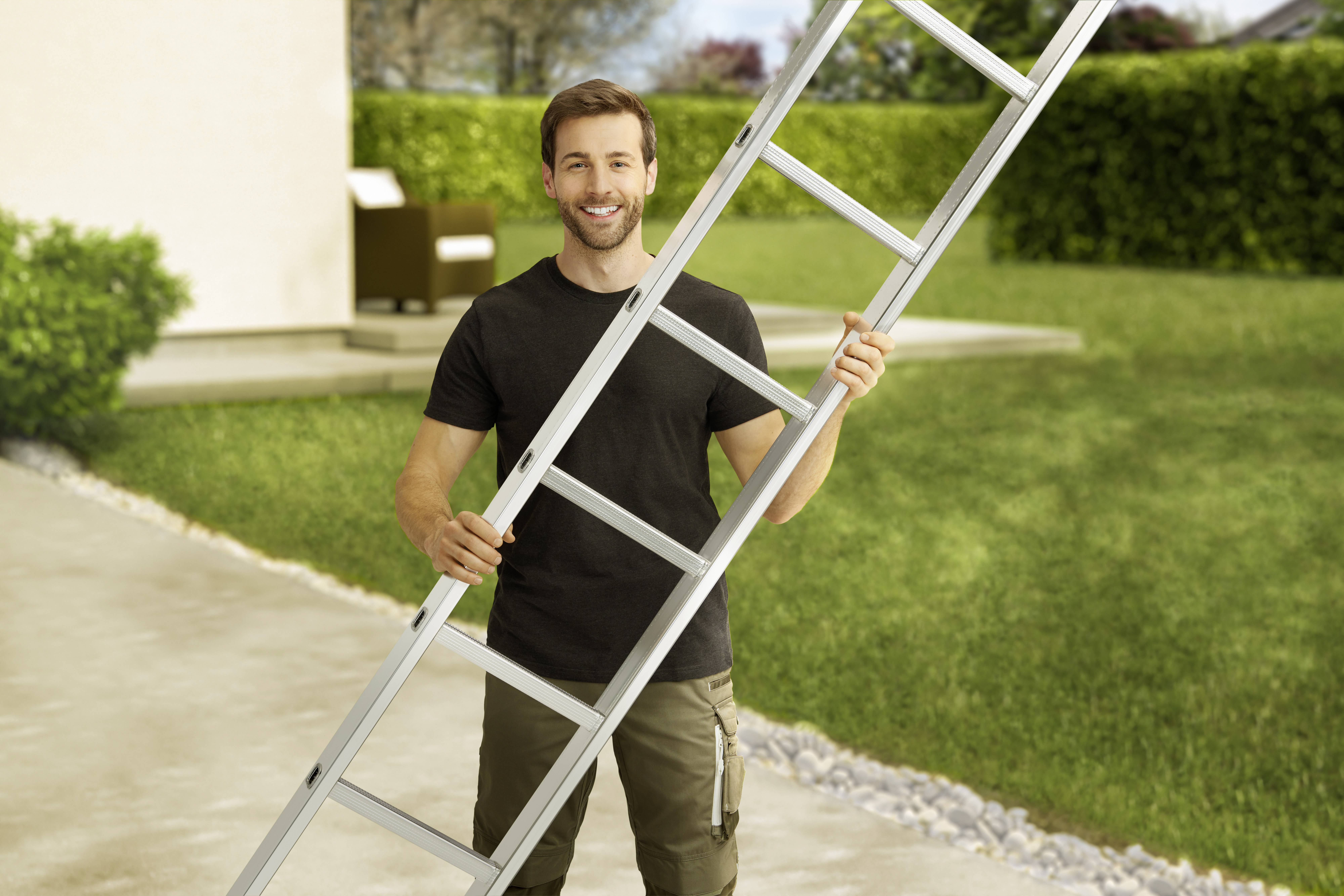 Ein Mann steht lächelnd in einem Garten und hält eine Aluminiumleiter. Im Hintergrund sind eine Terrasse und viel Grün zu sehen.