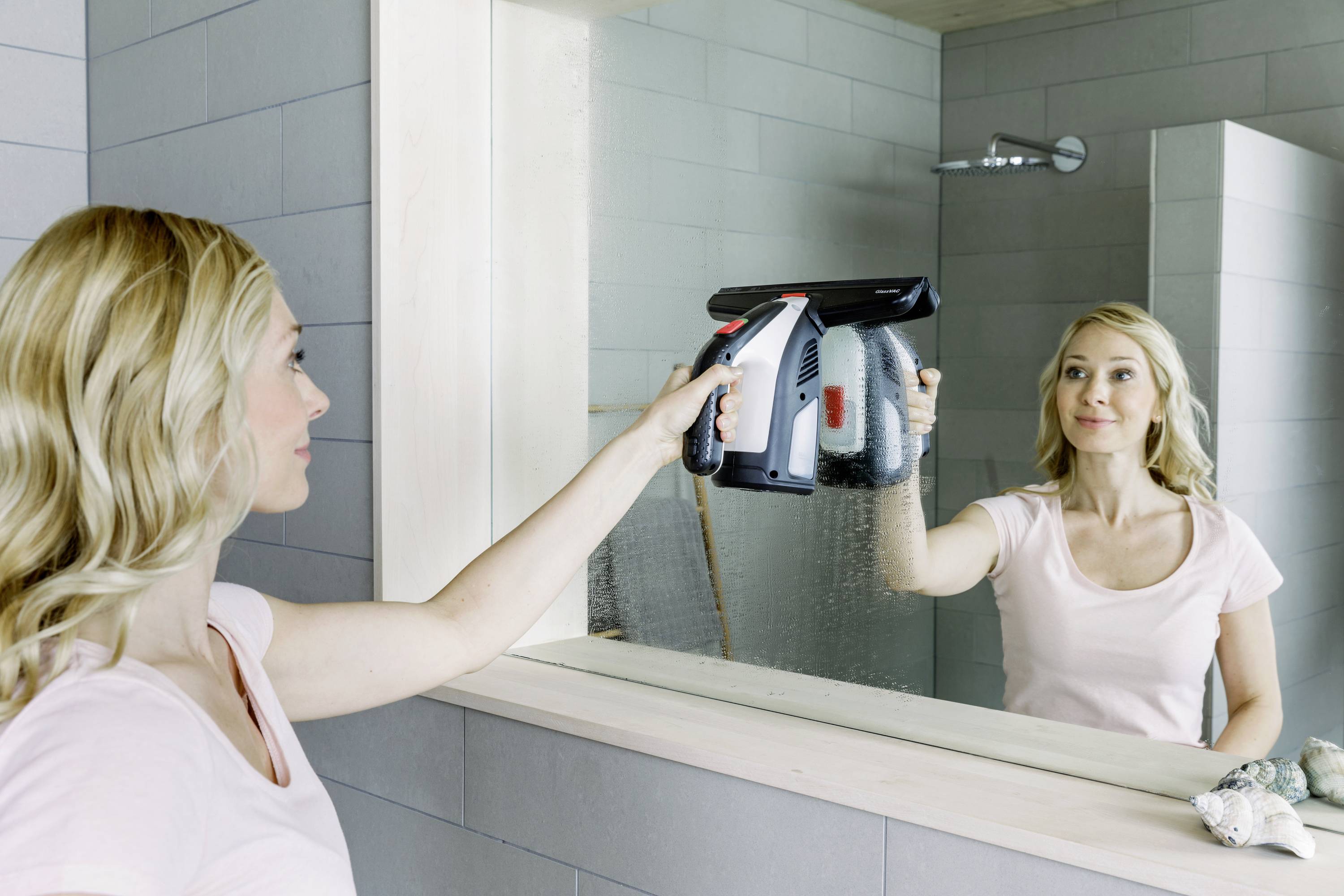 Eine Frau reinigt mit einem elektrischen Fensterreiniger einen Spiegel im Badezimmer. Sie trägt ein rosa T-Shirt und schaut konzentriert.