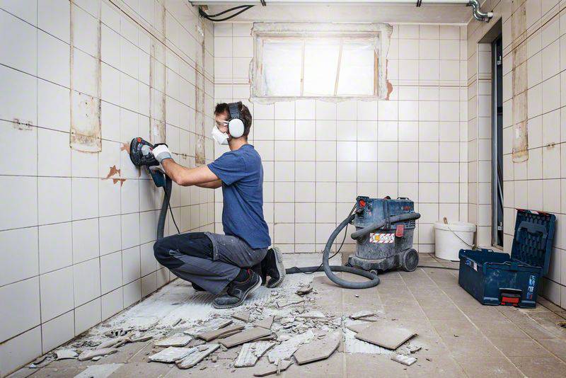 Ein Handwerker arbeitet in einem gefliesten Raum mit einer Schleifmaschine an der Wand, während Schutt den Boden bedeckt.