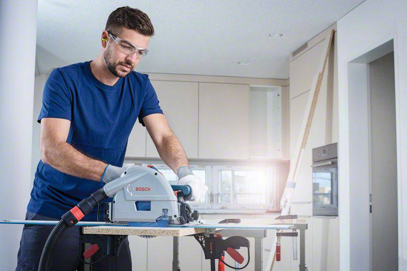 Ein Mann mit Schutzbrille und Gehörschutz arbeitet mit einer Kreissäge an einem Stück Holz in einer modernen Küche.