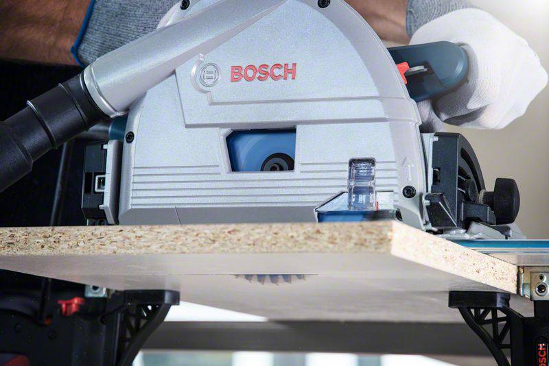 Elektrische Kreissäge von Bosch schneidet eine Holzplatte auf einem Tisch. Eine Person hält das Werkzeug, das an einen Staubsauger angeschlossen ist.