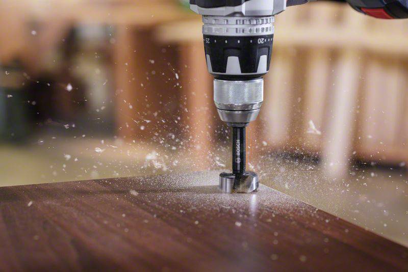 Bohrer durchbohrt ein Holzstück, Holzspäne fliegen. Arbeitsplatz im Hintergrund unscharf, Fokus auf die Präzision der Bearbeitung.