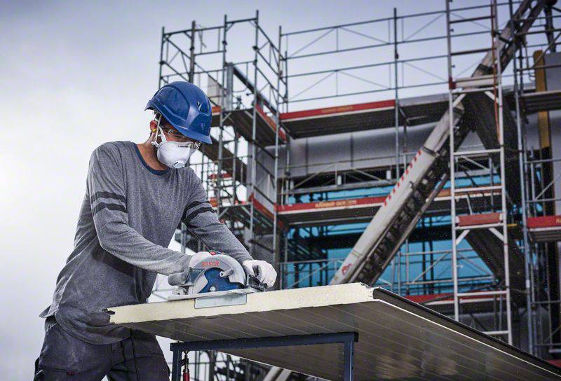 Ein Mann mit Schutzhelm und Maske schneidet mit einer Säge ein Metallblech, vor einem Gerüst auf einer Baustelle.