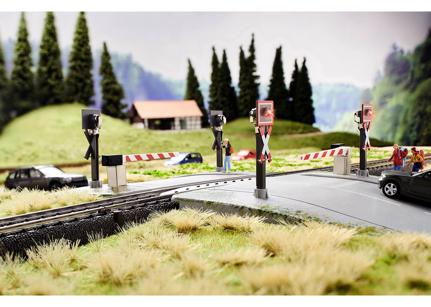 Eine Modelleisenbahnszene zeigt einen Bahnübergang mit geschlossenem Schranken und blinkenden Warnleuchten, Autos warten. Im Hintergrund sind Bäume und ein Haus.