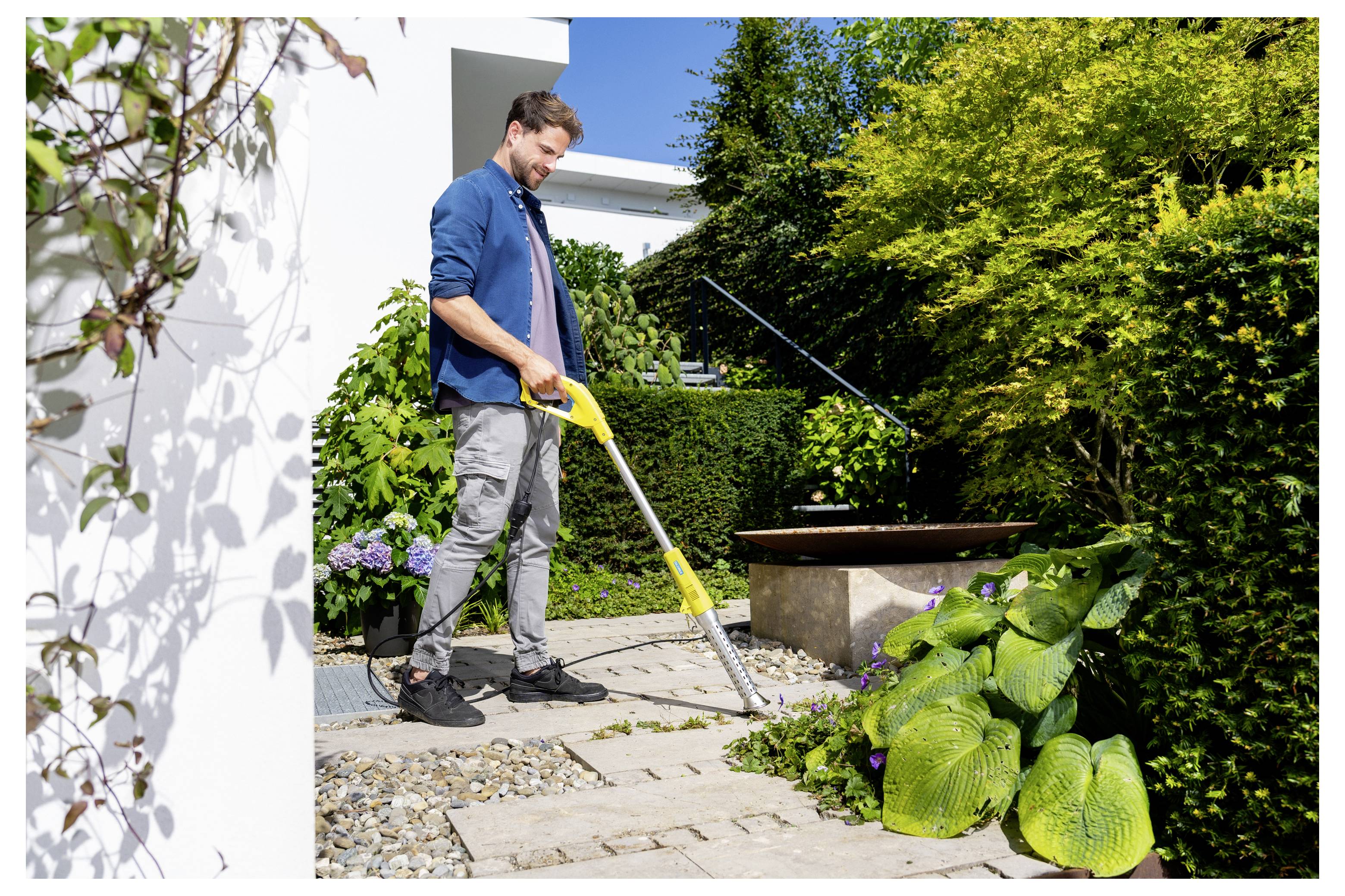 Ein Mann benutzt einen gelben Laubgebläse in einem angelegten Garten neben einer Terrasse und einem Teich und räumt Schutt in der Nähe von Sträuchern und Blumen weg.