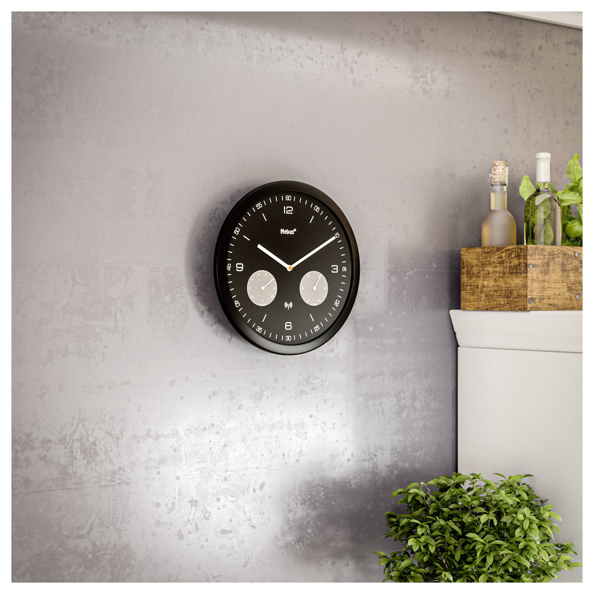 Eine moderne schwarze Wanduhr mit zwei Zeitzonen hängt an einer strukturierten grauen Wand; in der Nähe befinden sich grüne Pflanzen und eine Holzbox mit Flaschen.