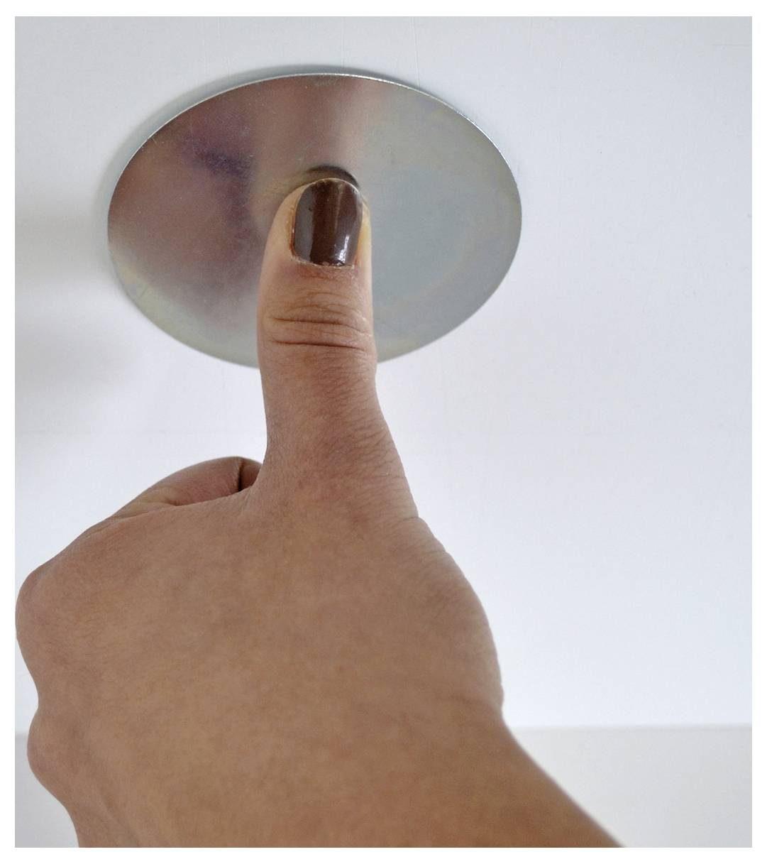 Eine Hand mit einem bemalten Daumennagel drückt einen runden metallischen Knopf oder eine Deckenleuchte gegen einen schlichten weißen Hintergrund.