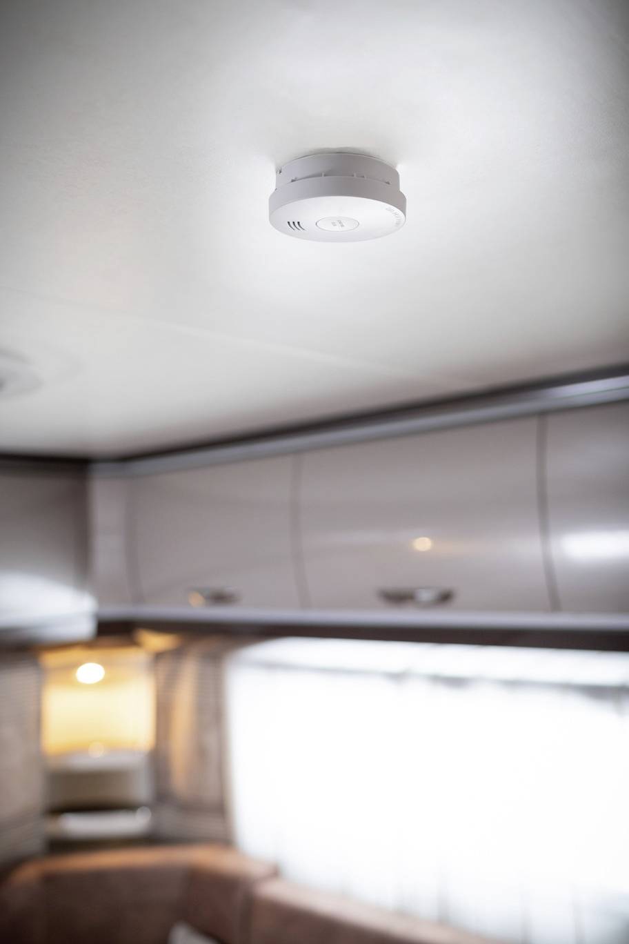 Rauchmelder an der Zimmerdecke eines Wohnwagens, umgeben von Schränken und einem beleuchteten Fenster im Hintergrund.