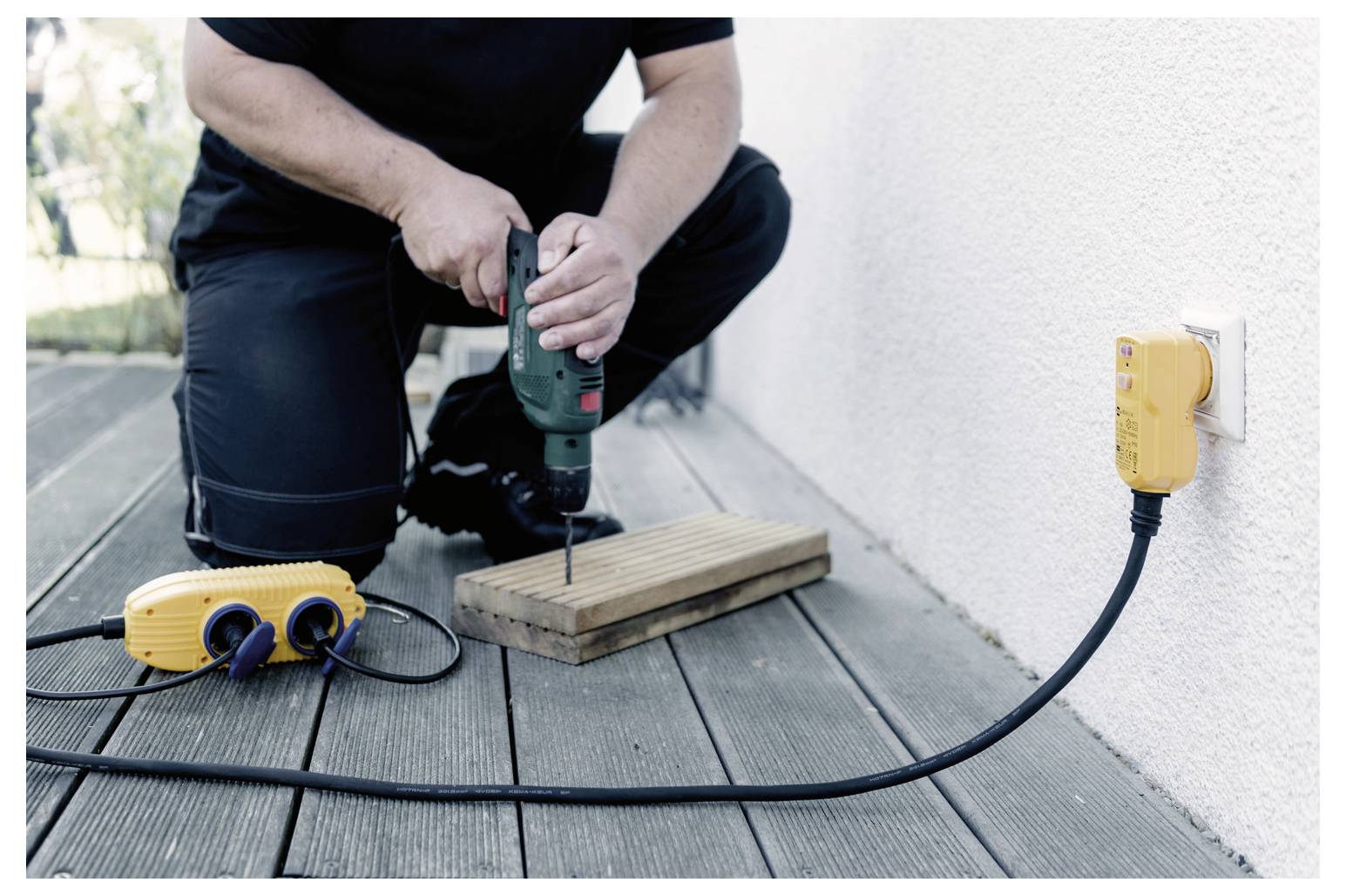 Eine Person kniet auf einem Holzdeck und bohrt in ein Holzbrett. Ein gelbes Verlängerungskabel und eine Steckdose sind in eine Wandsteckdose in der Nähe eingesteckt.