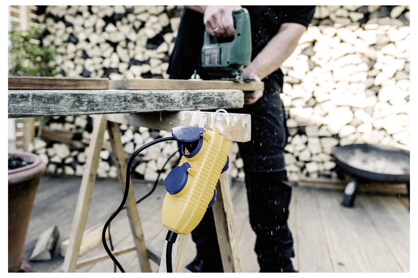 Person, die einen grünen Elektroschleiffer auf einem Holzbrett verwendet, mit einer gelben Verlängerungskabelrolle, die von der Werkbank in einer Werkstatt hängt.