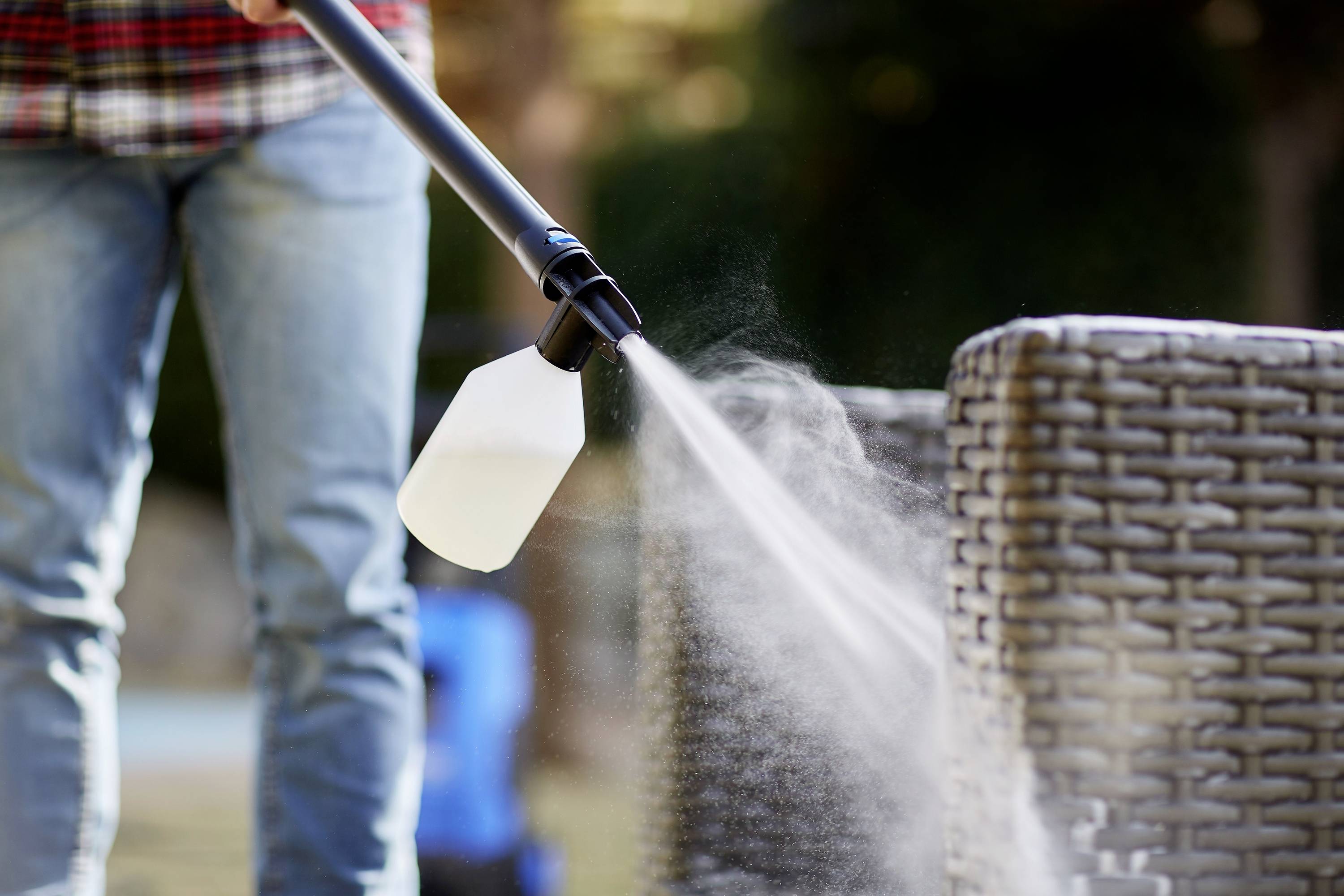 Eine Person reinigt ein Rattan-Möbelstück im Freien mit einem Hochdruckreiniger. Dampf oder Sprühnebel ist deutlich sichtbar.