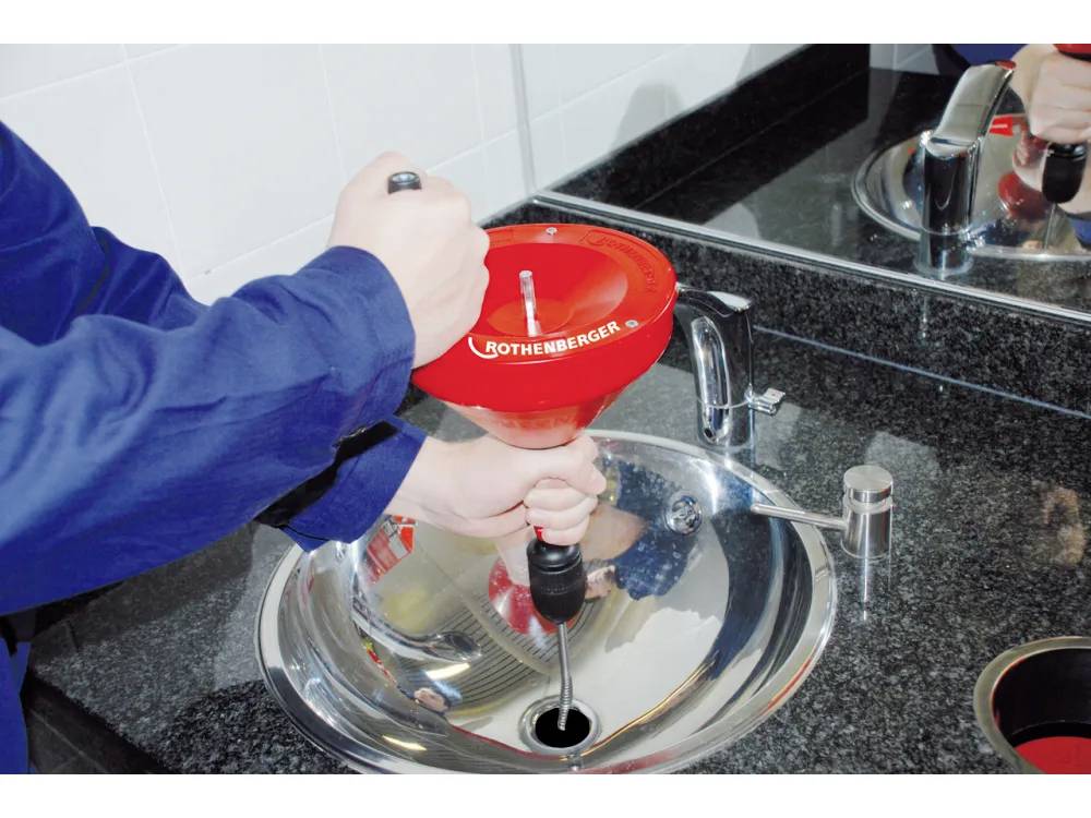 Person, die einen roten Rohrreinigungsbohrer an einem Waschbecken verwendet, um eine Verstopfung zu beseitigen. Das Waschbecken befindet sich neben einem reflektierenden Spiegel.