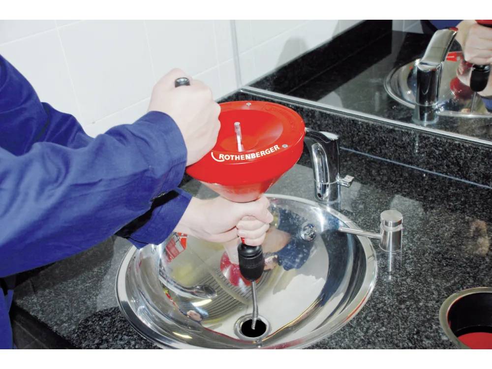 Eine Person verwendet einen roten Kolben mit Griff in einem silbernen Waschbecken, um es zu entklotzen. Der Hintergrund zeigt eine schwarze Arbeitsplatte und eine geflieste Wand.