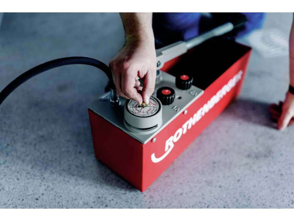 Eine Person, die einen Druckmesser an einem roten hydraulischen Werkzeug einstellt, das auf einer gesprenkelte grauen Oberfläche steht. Das Werkzeug hat einen Schlauch, der daran angeschlossen ist.