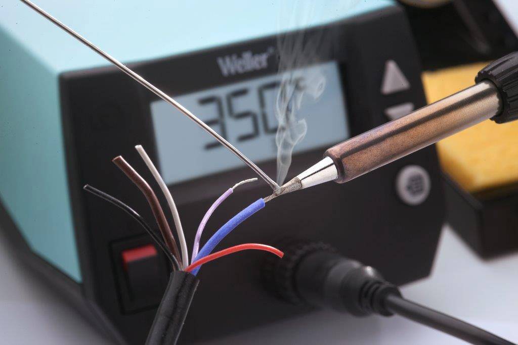 Ein Lötkolben erhitzt Drähte neben einer Anzeige mit der Temperatur '250°C'. Rauch steigt auf. Fokus auf Löttechnik.