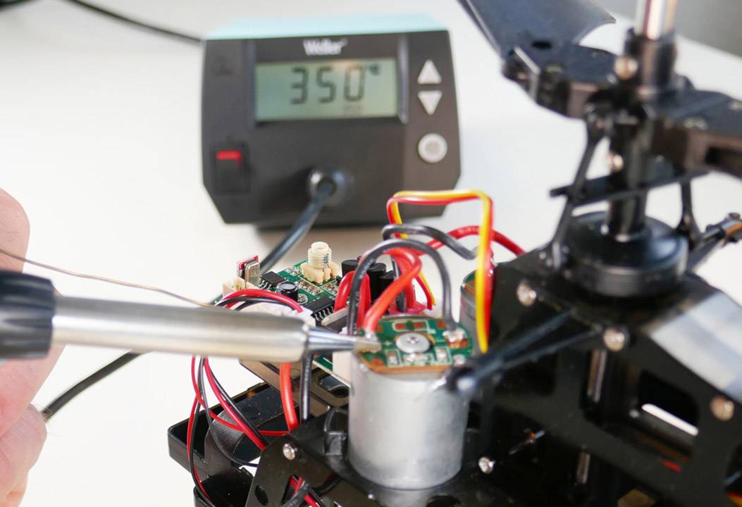 Eine Person lötet an einer Platine mit einem Lötkolben. Im Hintergrund zeigt ein digitales Thermometer 350 Grad Celsius an.