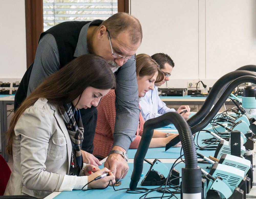 Ein Lehrer hilft einer Studentin bei einer Lötübung in einem Elektroniklabor, während andere Studierende konzentriert an ihren Projekten arbeiten.