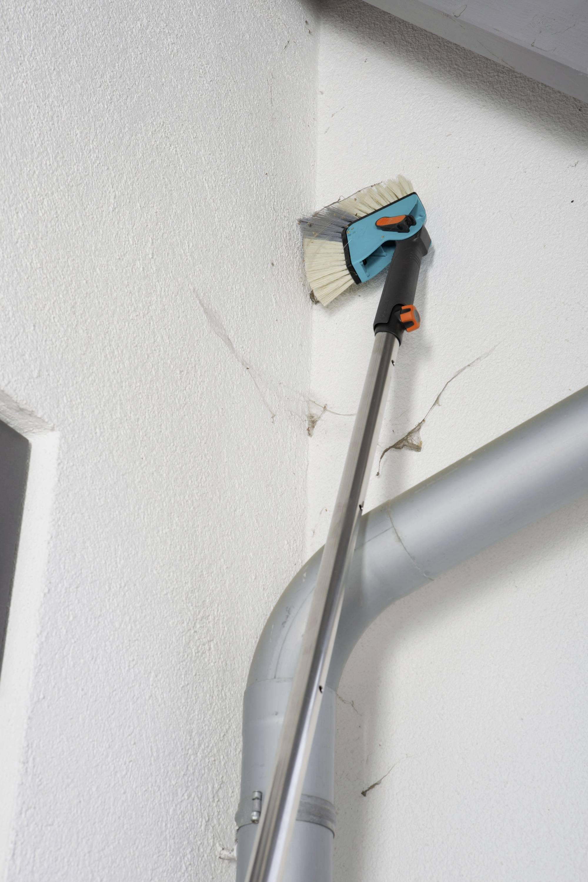 Eine Person reinigt mit einem Teleskopbesen Spinnweben an der Decke eines Zimmers, neben einem grauen Rohr.
