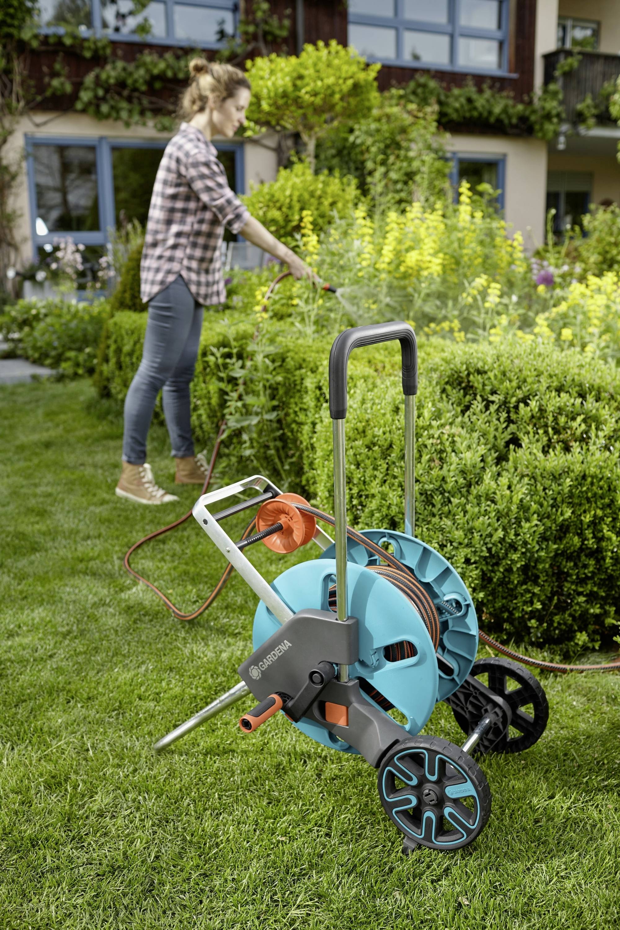Eine Person gießt einen Garten mit einem Schlauch, der an einem blauen Schlauchwagen befestigt ist. Der Garten ist grün und blüht.