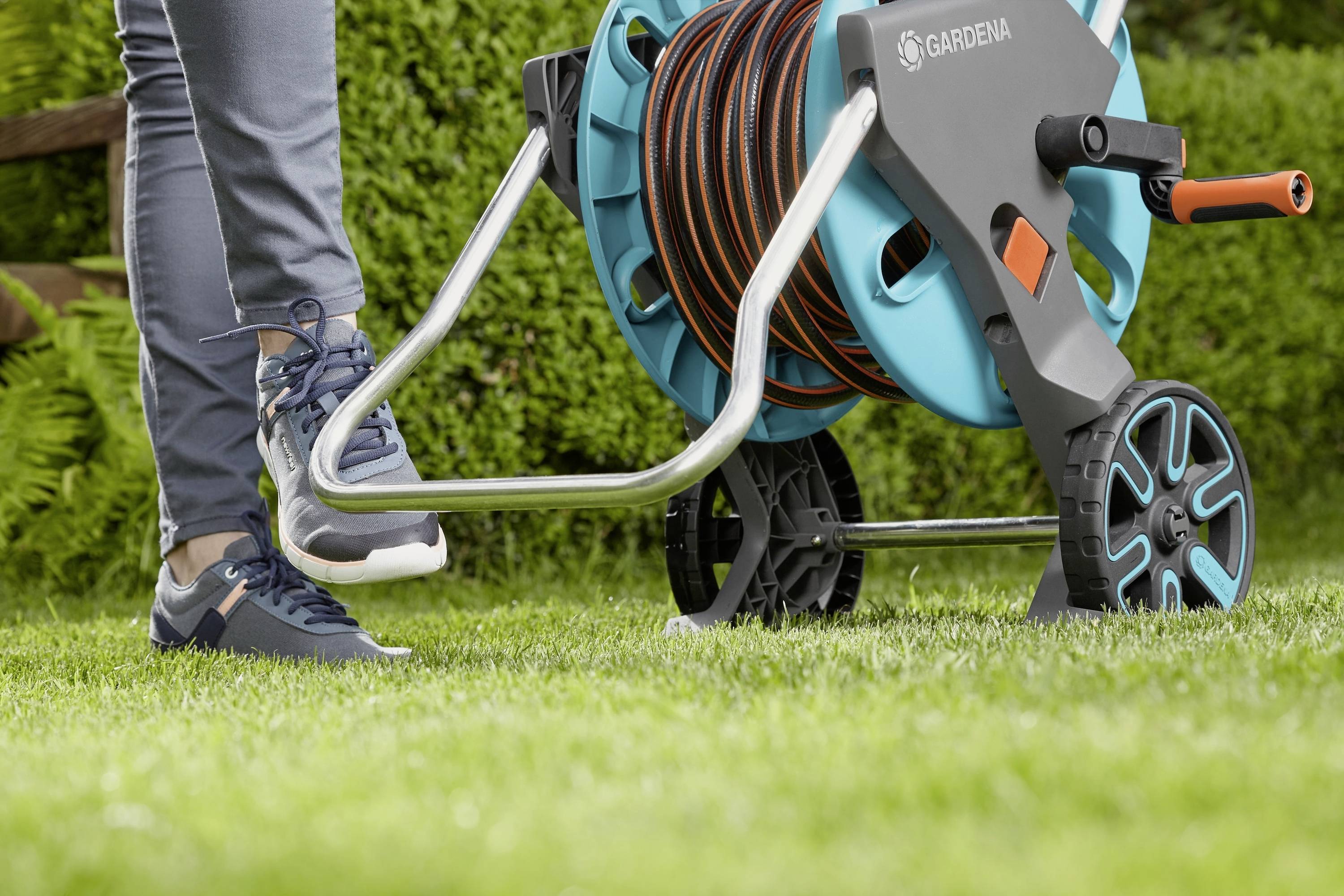 Person tritt auf den Fußhebel eines Schlauchwagens auf einer Rasenfläche. Der Wagen hat einen aufgewickelten Gartenschlauch.