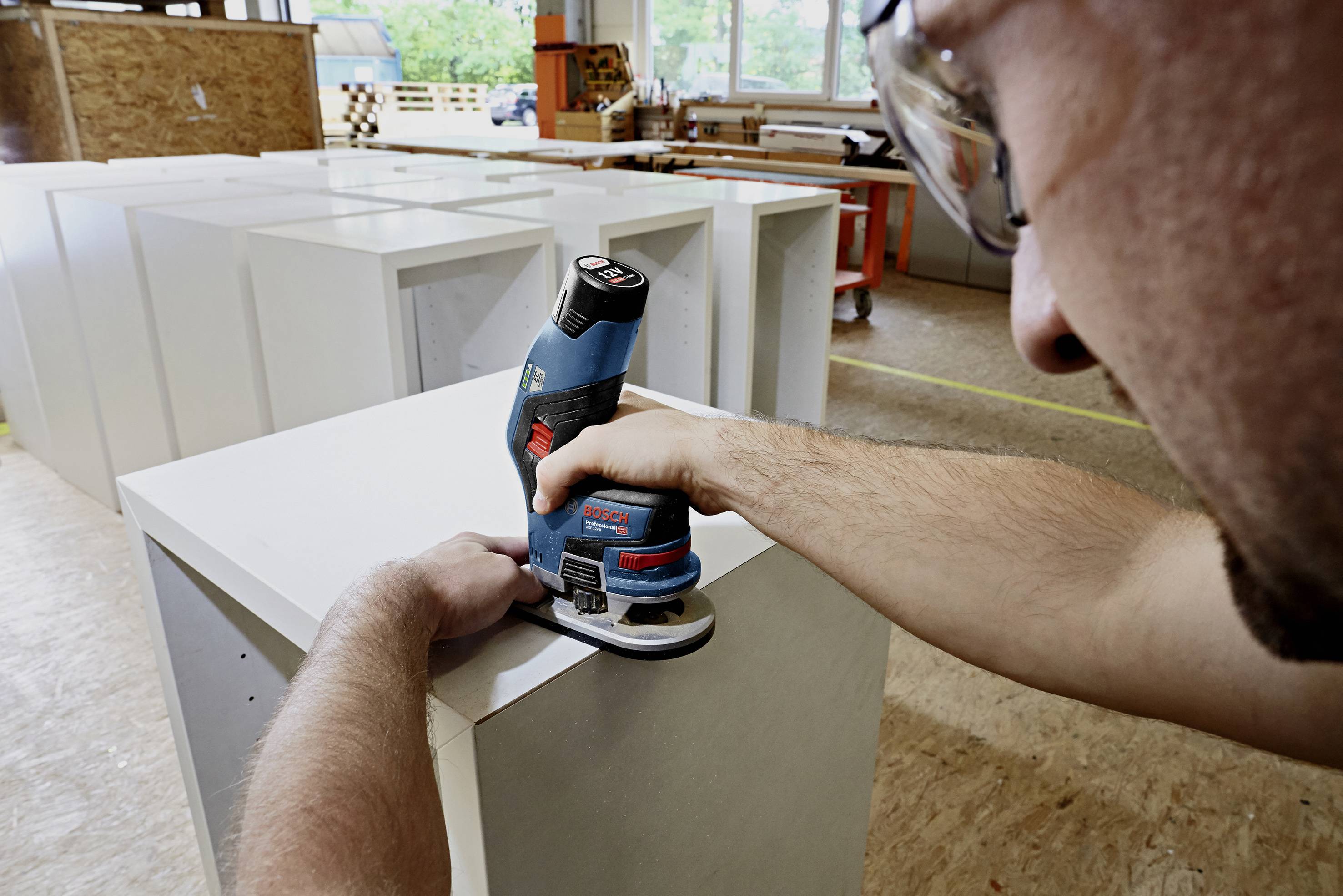 Eine Person schleift mit einem Elektrowerkzeug sorgfältig die Kanten eines großen weißen Möbelstücks in einer Werkstatt.