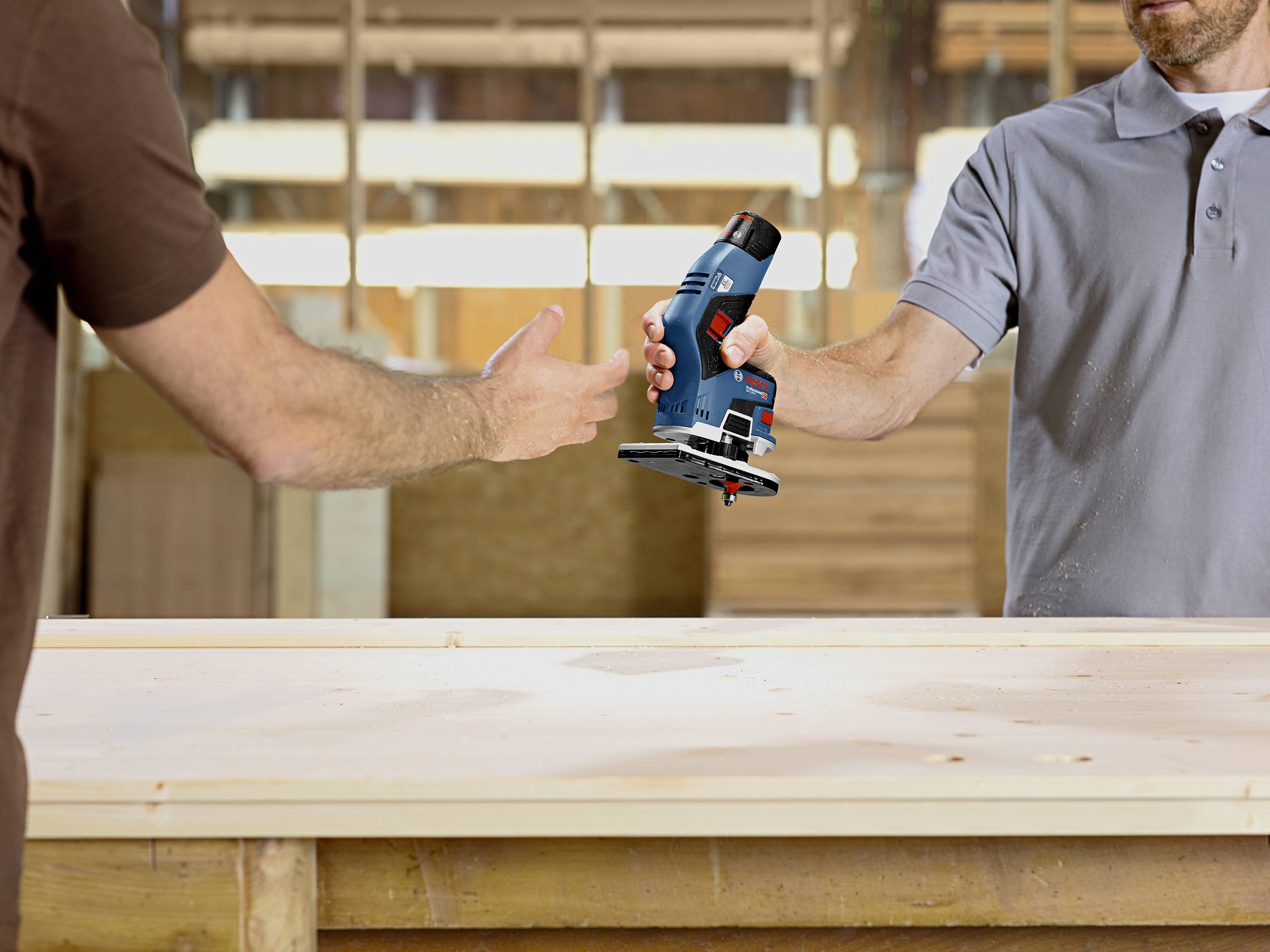 Zwei Personen übergeben sich eine elektrische Fräse über eine Werkbank in einer Werkstattumgebung.
