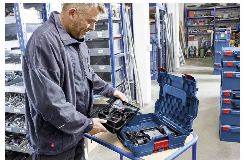 Ein Arbeiter in einem Lagerhaus inspiziert ein kabelloses Elektrowerkzeug und ein Batteriesatz in einem offenen blauen Transportkoffer auf einem Wagen.
