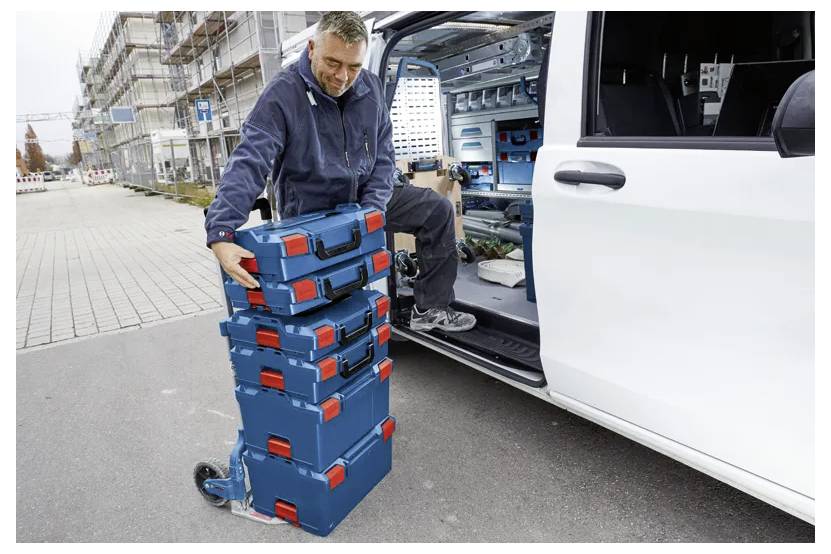 Eine Person stapelt einen Turm aus blauen und roten Werkzeugkästen auf einem Rollwagen neben einer offenen Van-Tür, was eine Arbeitssituation oder Werkzeugorganisation andeutet.