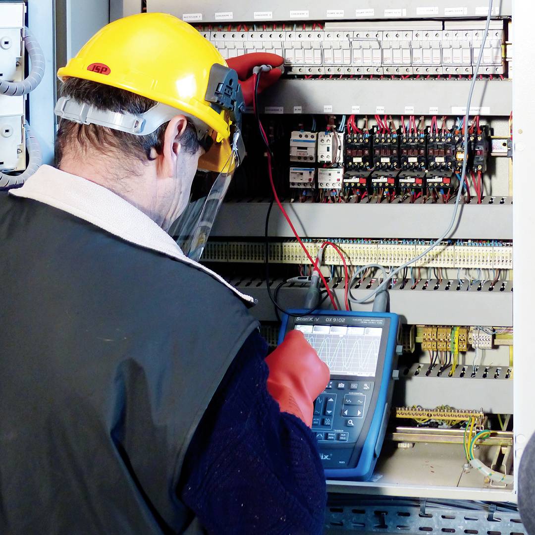 Ein Techniker mit Schutzhelm und Visier arbeitet an einem offenen Schaltschrank, während er ein Multimeter verwendet.