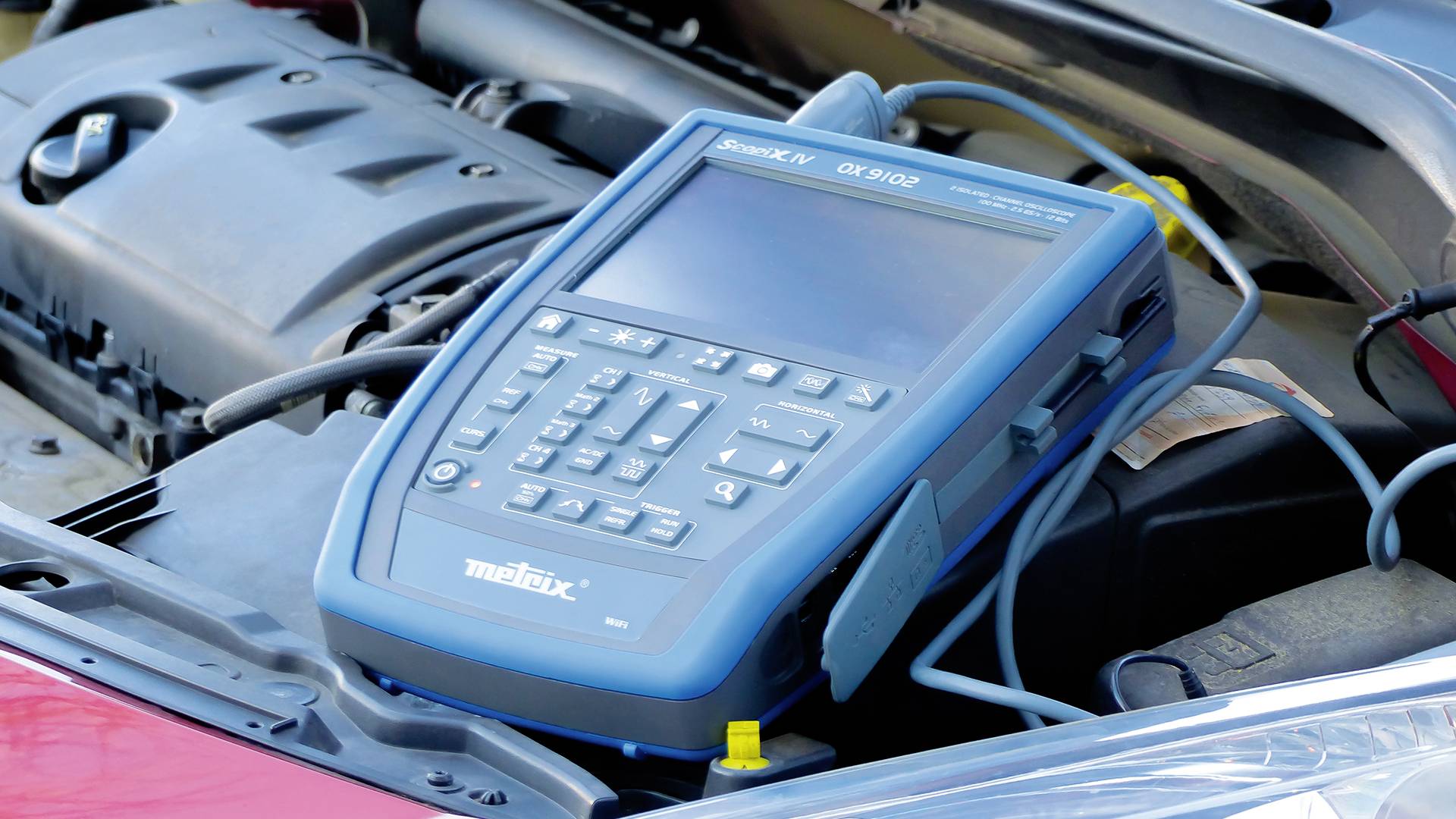Ein blaues Diagnosegerät ist unter der geöffneten Motorhaube eines Autos platziert, um den Fahrzeugstatus zu überprüfen.