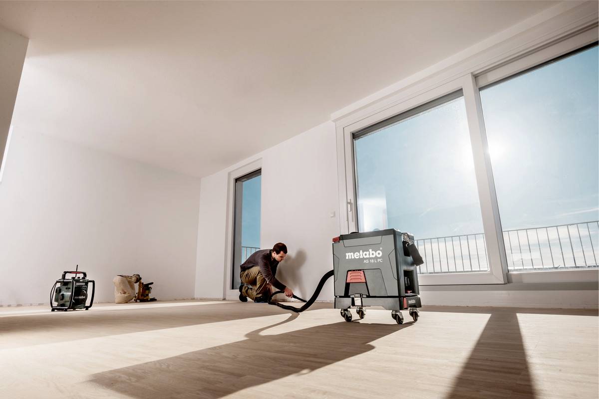 Eine Person verlegt einen Holzboden in einem hellen Raum mit großen Fenstern. Ein Staubsauger von 'Metabo' steht neben der Person.