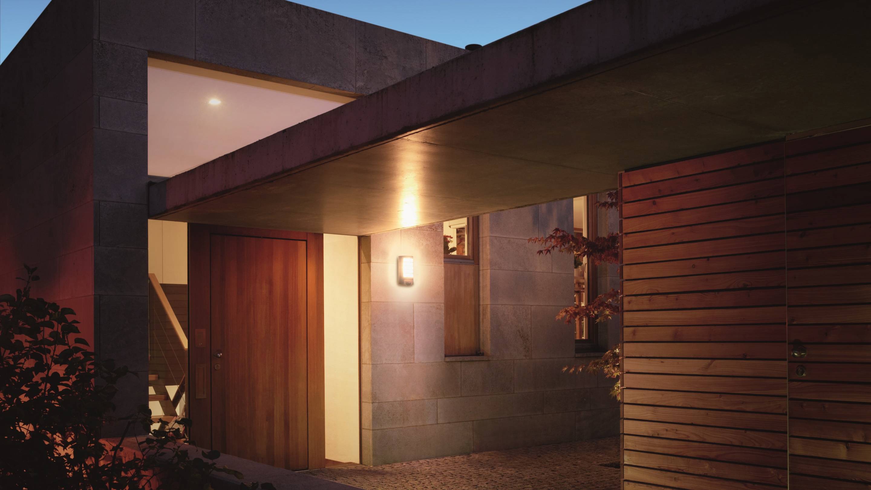 Modernes Haus bei Dämmerung, warm beleuchtet. Holz- und Steinmaterialien sind sichtbar. Vor der Tür strahlt ein Außenlicht.