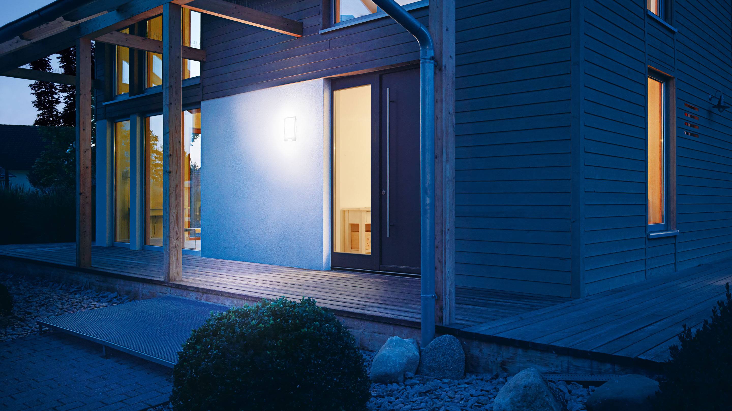 Ein modernes Haus bei Nacht, beleuchtet von einer Wandleuchte neben der Haustür. Veranda und Gartenpflanzen im Vordergrund.