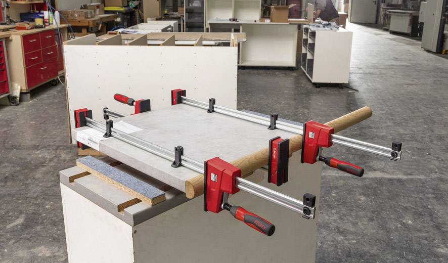 Eine Werkbank mit mehreren Klammern, die ein Holzstück fixieren. Im Hintergrund sind weitere Werkzeuge und Möbelstücke in einer Werkstatt sichtbar.