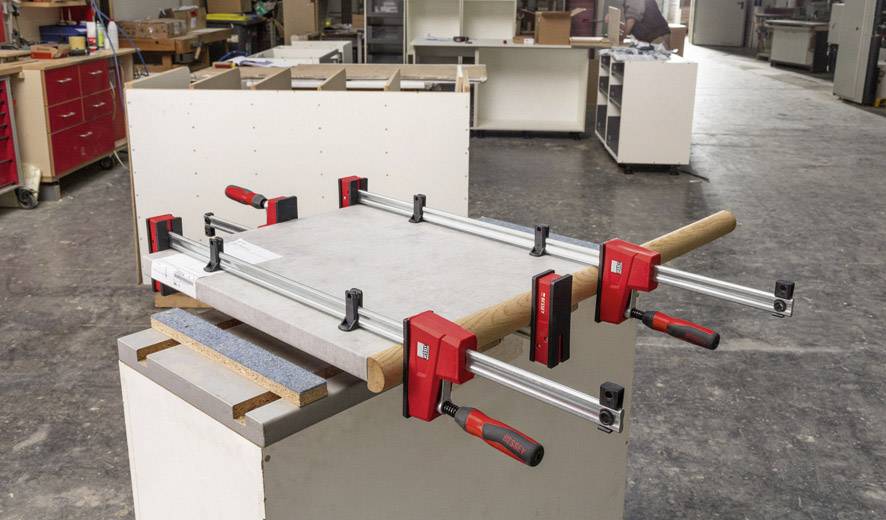 In einer Werkstatt wird eine Holzplatte mit mehreren Schraubzwingen auf einer Werkbank fixiert. Im Hintergrund sind Regale zu sehen.