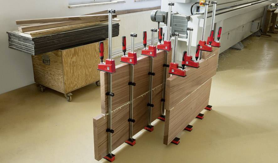 Holzplatten werden in einer Werkstatt mit mehreren Zwingen in Position gehalten. Im Hintergrund ist eine Holzbearbeitungsmaschine zu sehen.