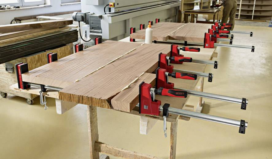 Holzbretter werden mit roten Schraubzwingen auf einem Tisch festgeklemmt, um sie zum Verleimen zu fixieren. Eine Werkstattumgebung.