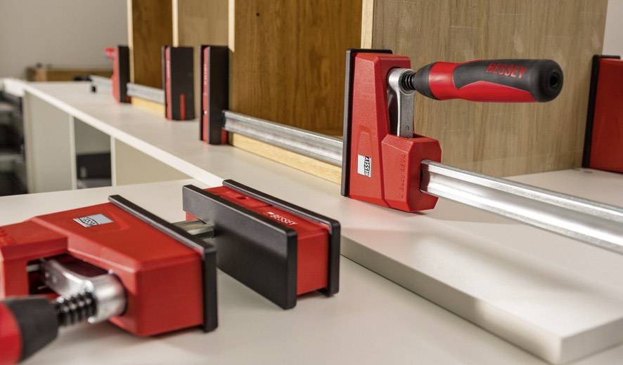 Mehrere rote Zwingen halten Holzplatten zusammen auf einer weißen Arbeitsfläche, während im Hintergrund ein Regal montiert wird.