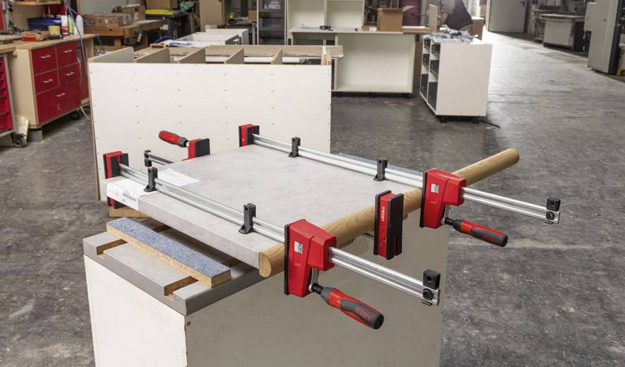 Mehrere große rote Zwingen klemmen eine Holzplatte auf einem Tisch in einer Werkstatt. Im Hintergrund sind Regale und weitere Möbelteile erkennbar.