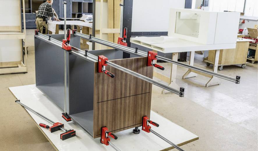 Ein Arbeiter in einer Werkstatt montiert ein großes Holzmöbelstück mit mehreren Schraubzwingen, um die Teile zusammenzuhalten.